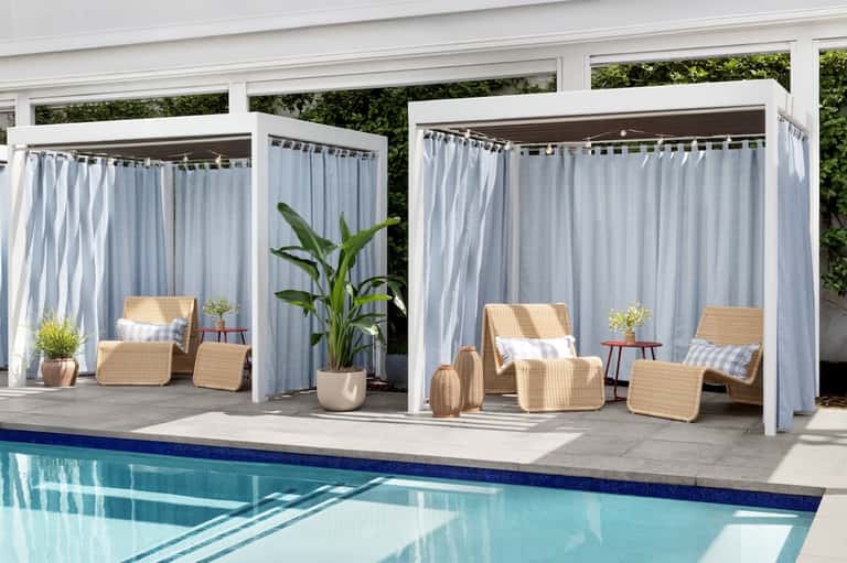 Poolside cabanas with white curtains, woven lounge chairs, potted plants, and blue swimming pool