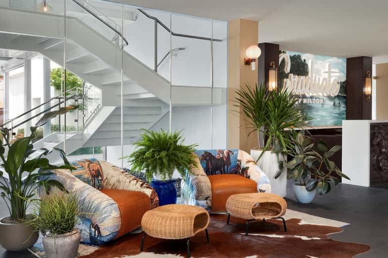 Modern hotel lobby with curved orange sofa, wicker chairs, plants, and staircase with glass railings