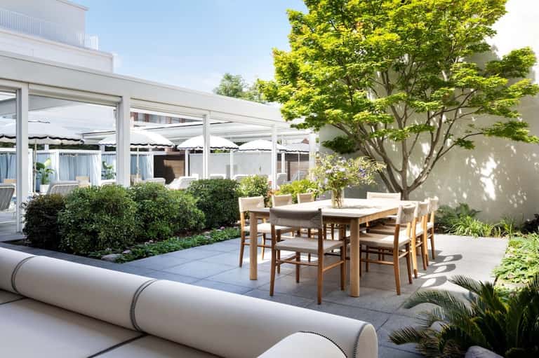 Modern terrace with dining table, wooden chairs, large trees, and architectural pergola structure