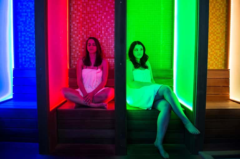Two women sitting in colorful neon-lit spa pods with blue, red, and green ambient lighting