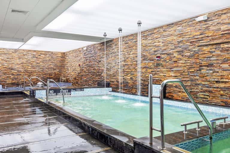 Indoor hot springs pool with green water, stone accent wall, glass roof, and metal railings
