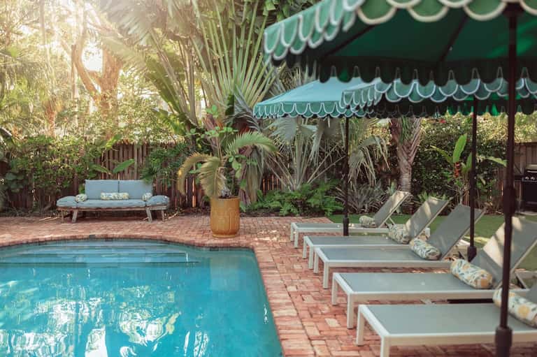 Residential pool with brick patio, lounge chairs under green awning, palm trees, and garden seating
