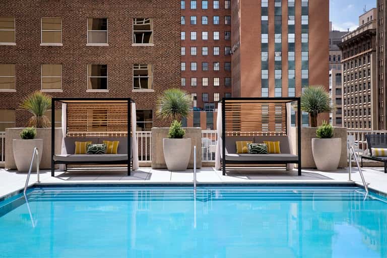 Urban rooftop pool with bright blue water, black cabanas with yellow pillows, and downtown city buildings