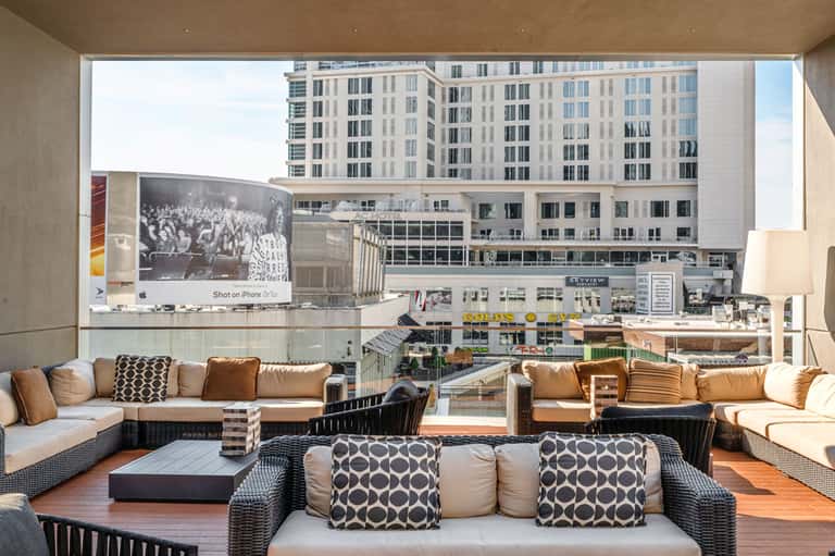 Modern rooftop lounge with tan wicker seating, geometric pillows, and city skyline views through floor-to-ceiling windows