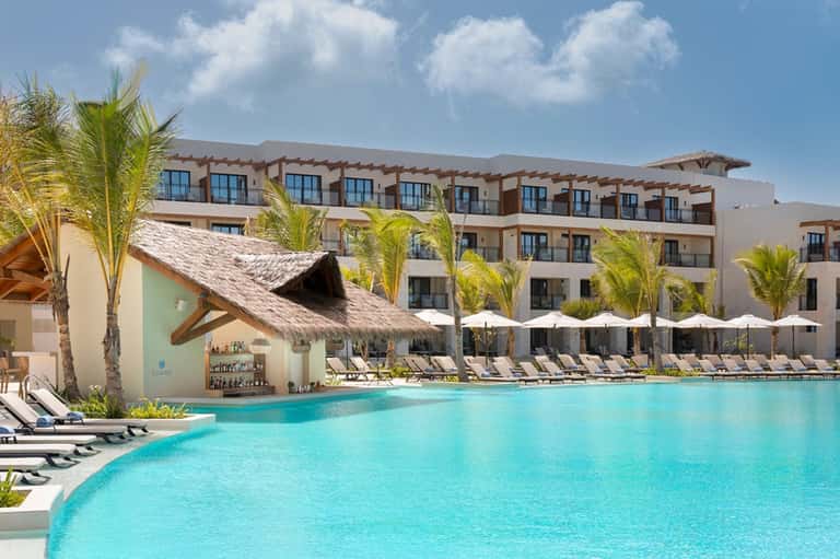 Tropical resort pool with thatched-roof bar, palm trees, white umbrellas, and loungers