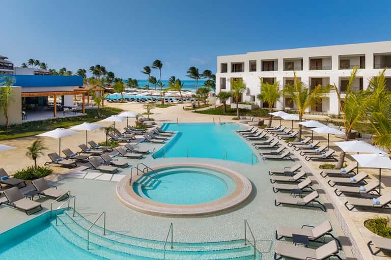Resort courtyard with turquoise pool, spa tub, white building, and ocean view in background