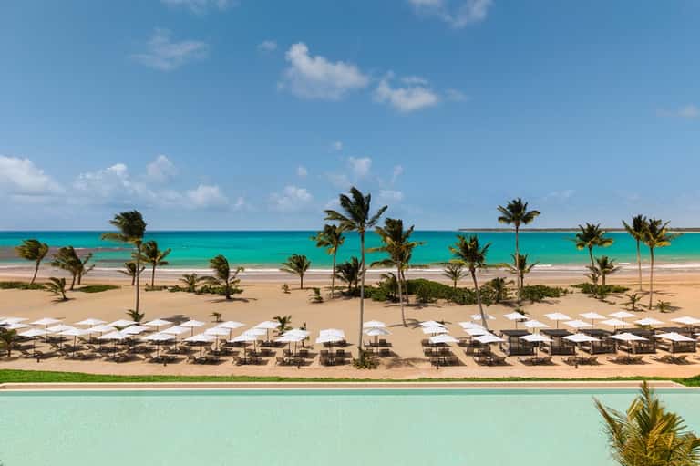 Aerial view of pristine beach with palm trees, white umbrellas, turquoise pool, and Caribbean ocean