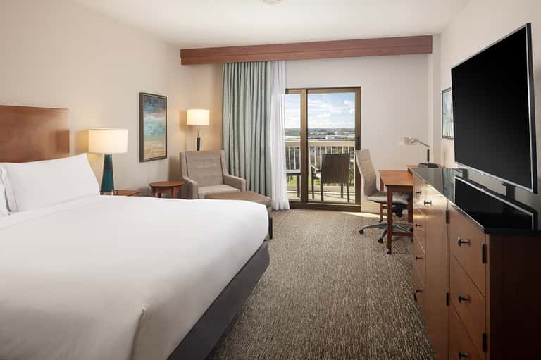 Hotel bedroom with queen bed, balcony overlooking waterfront, sitting area, TV, and modern furnishings
