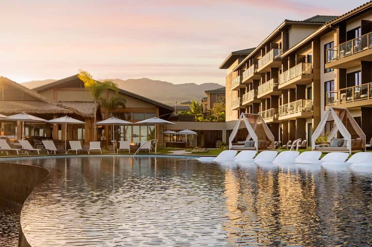 Luxury resort pool at sunset with mountain views, white cabanas, lounge chairs, and multi-story hotel building