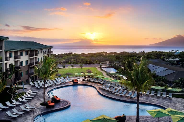 Scenic resort pool at sunset with mountain views, lounge chairs, palm trees, and curved infinity pool