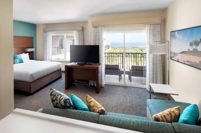 Modern hotel suite with queen bed, flat-screen TV, turquoise accents, and ocean view balcony