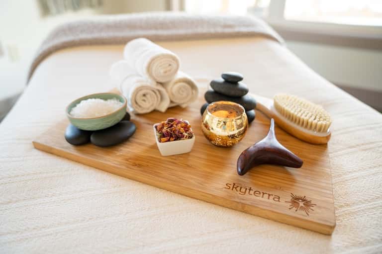 Spa amenities display with rolled towels, salt, stones, brush, and branded wooden tray on bed