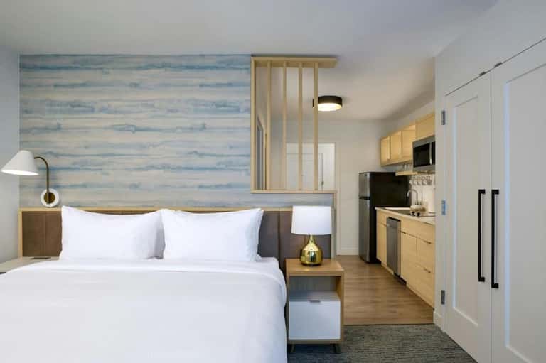 Compact hotel room with queen bed, kitchenette, wood accents, and blue wallpaper