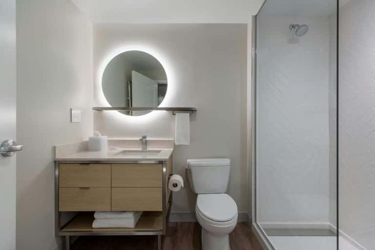 Contemporary bathroom with illuminated circular mirror, wooden vanity, and walk-in shower