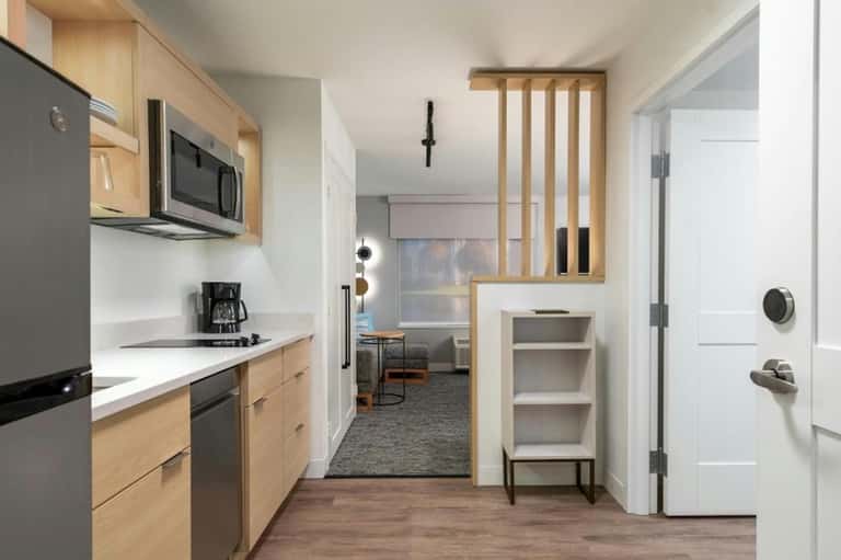Modern hotel suite kitchen with wood cabinetry, stainless appliances, and living area beyond