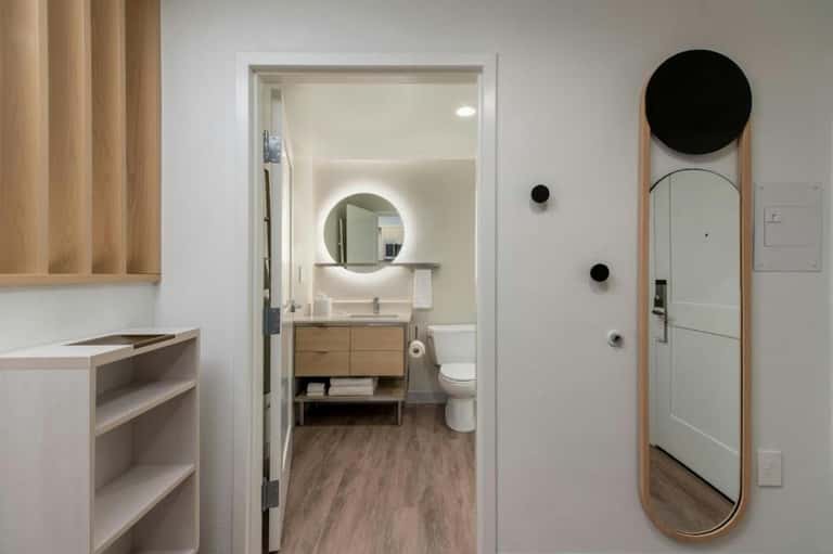 Modern bathroom with glowing circular mirror, wooden vanity, open doorway, and minimalist design