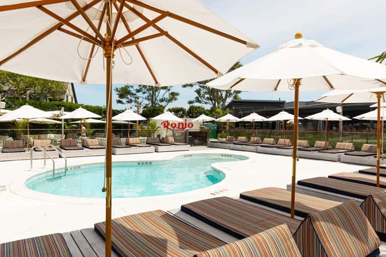 Resort pool area with lounge chairs, wooden umbrellas, white shade structures, and manicured landscaping