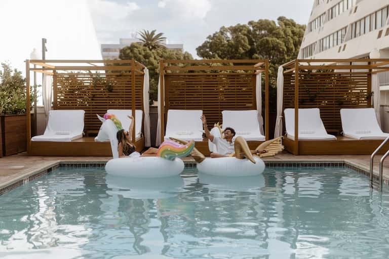 Guests relaxing in pool with private cabanas featuring white lounges and wooden pergola structures