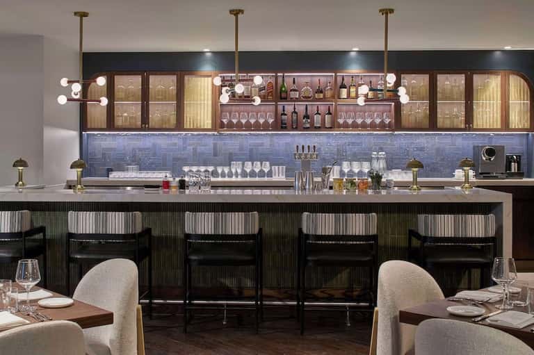Upscale bar and restaurant with blue tile backsplash, illuminated shelving, modern pendant lights, and dining area