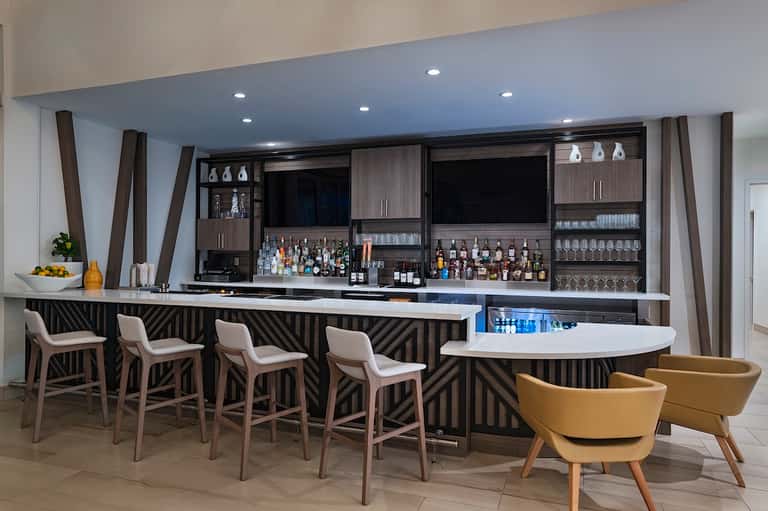 Modern hotel bar with dark geometric wood design, white bar seating, and liquor display