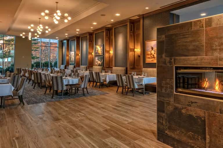 Upscale hotel dining room with fireplace, wood paneling, large windows, and tables set for fine dining with warm lighting