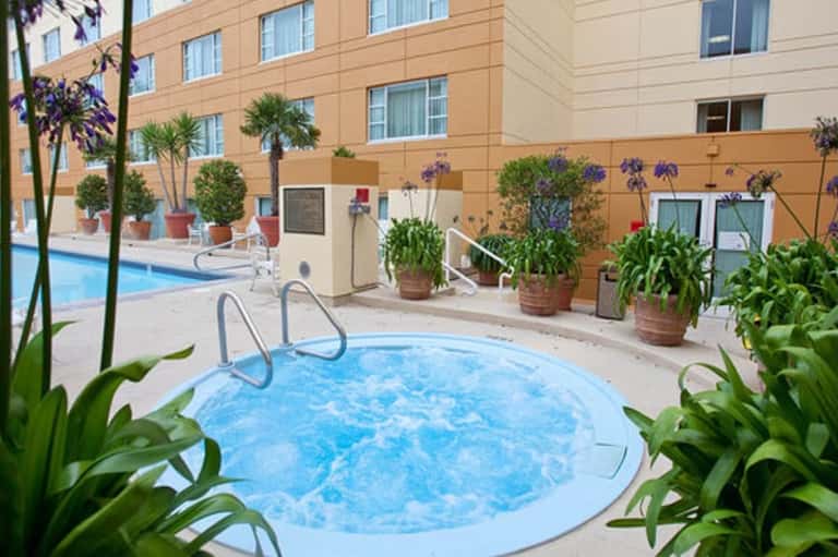 Hot tub surrounded by potted plants with resort building and larger pool visible in background courtyard