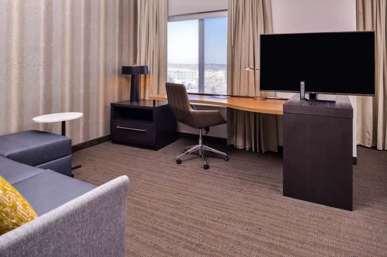 Hotel suite with dark wood desk, flat-screen TV, and city views through large windows with beige curtains