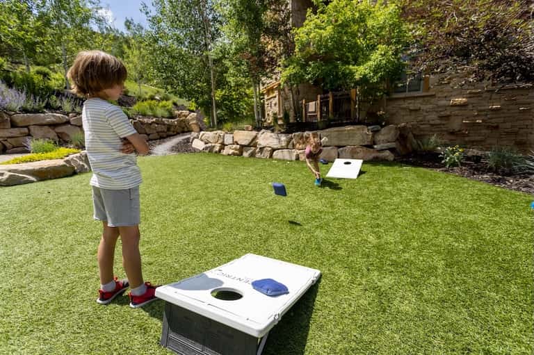 Child playing cornhole in a manicured lawn garden with stone walls and trees
