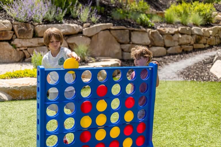 Two young children playing giant Connect Four game on a sunny lawn with stone wall backdrop
