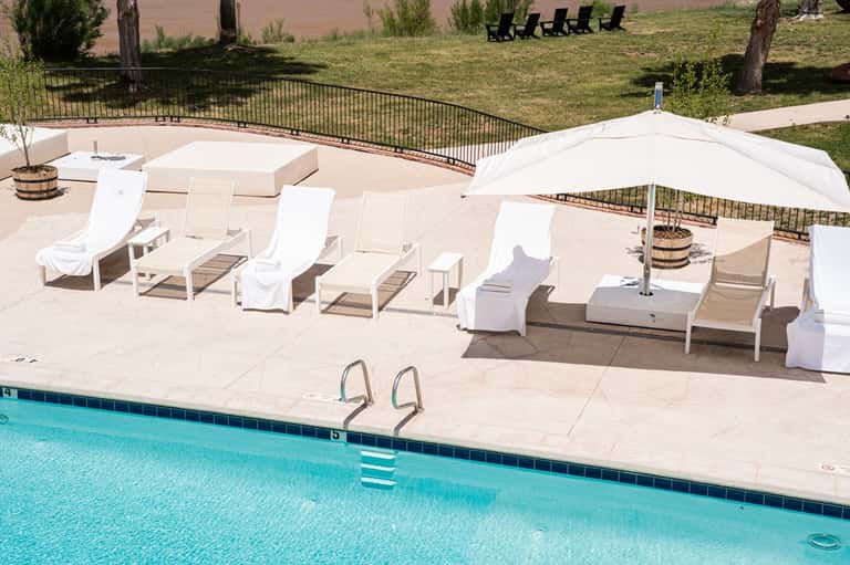 Resort pool deck with white lounge chairs, umbrellas, fire pit, and manicured lawn with trees