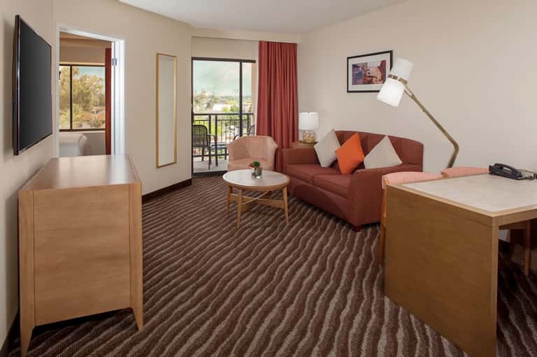 Hotel suite with balcony view, brown sofa, work desk, and television entertainment
