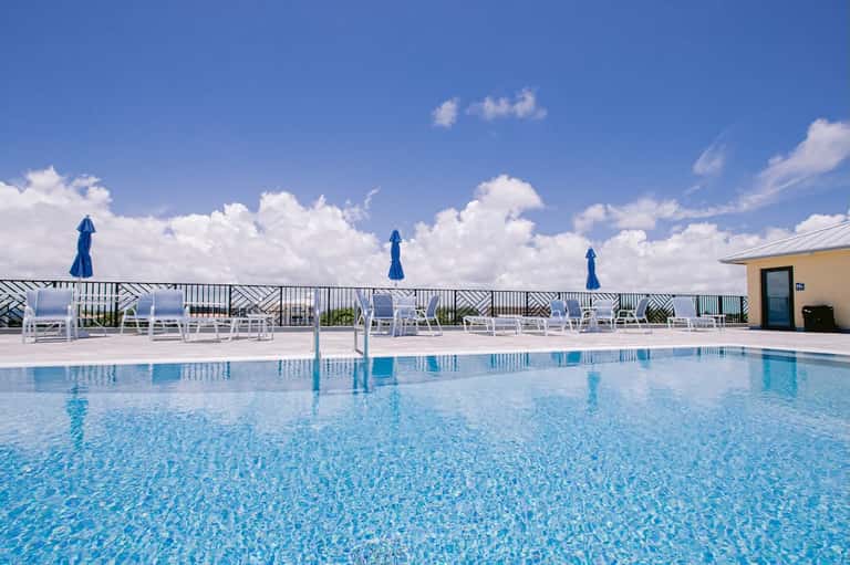 Resort pool with white loungers, blue umbrellas, and ocean views beyond railing