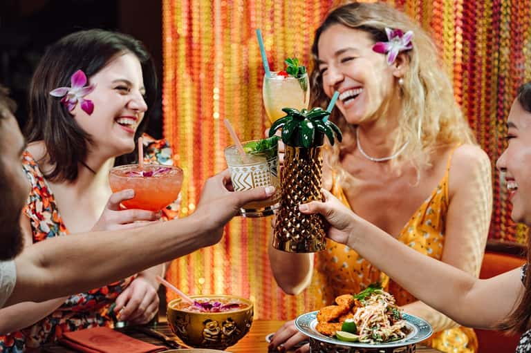 Women clinking tropical cocktails at vibrant dinner celebration with colorful beaded backdrop and Asian cuisine plating