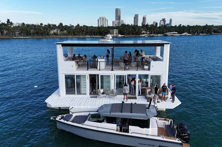 Luxury floating platform structure on blue water with guests dining, glass windows, and city skyline in background