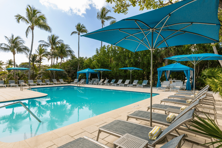 Resort pool with turquoise water, palm trees, blue umbrellas, and lounge seating