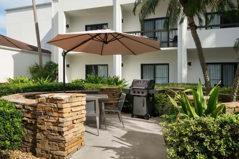 Outdoor patio with gas grill, stone fireplace, large umbrella, and modern resort building backdrop