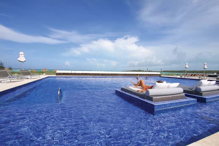 Rectangular pool with floating loungers, ocean and sky view, white umbrellas, and woman relaxing on white cushioned seating