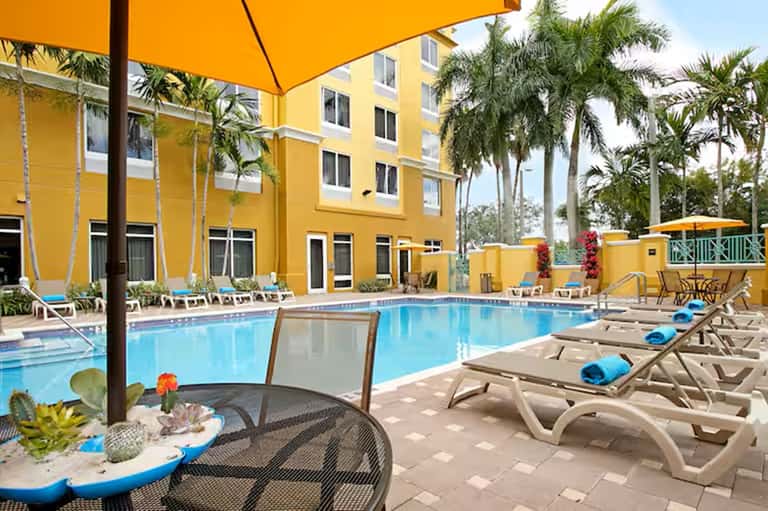 Resort pool with yellow building facade, palm trees, lounge chairs, umbrellas, and water features