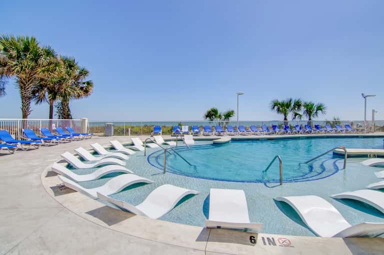 Curved swimming pool with white lounge chairs, blue lounge chairs in background, palm trees, and ocean view over railing