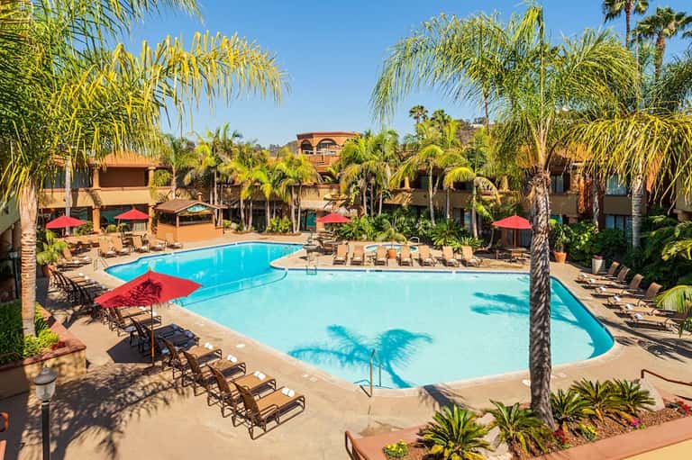 Resort pool area with multiple swimming pools, red umbrellas, palm trees, and lounge chairs