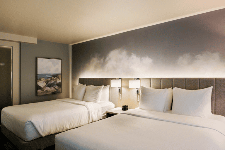 Hotel guest room with two queen beds, gray upholstered headboards, white linens, and beach artwork