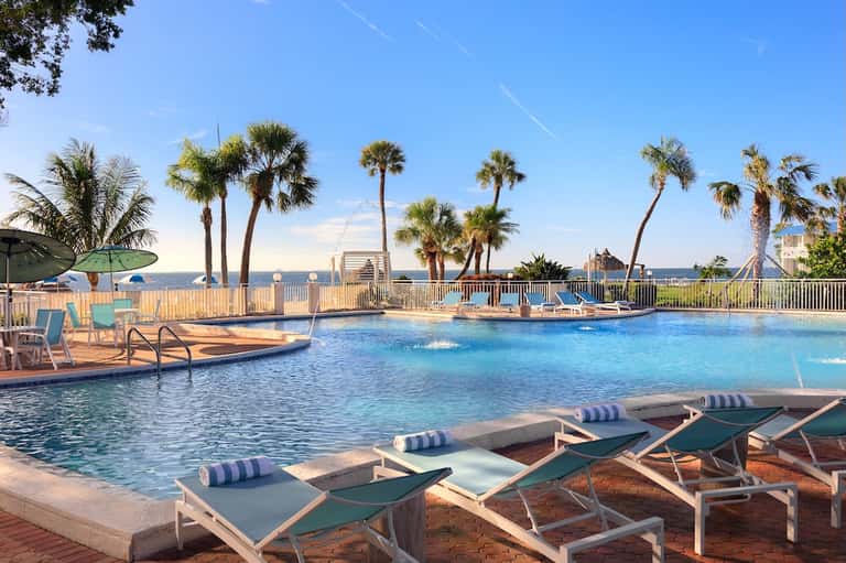 Resort outdoor pool area with lounge chairs, palm trees, ocean view, wooden deck, and clear blue water