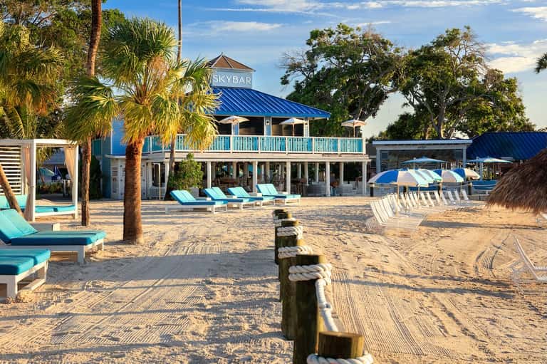 Beachfront skybar with blue metal roof, white railings, lounge chairs, and palm trees on sandy beach