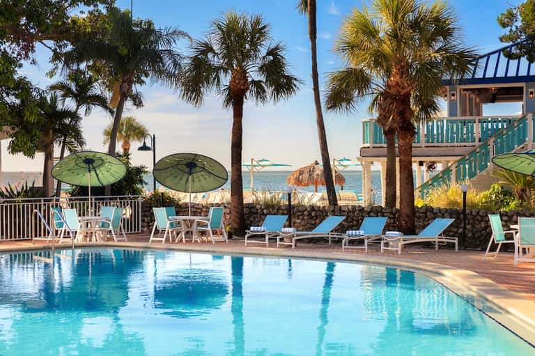 Resort pool with turquoise water, palm trees, green umbrellas, and ocean view backdrop