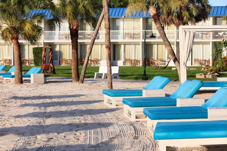 Beach resort with white beachfront building, blue lounge chairs on sand, and palm trees