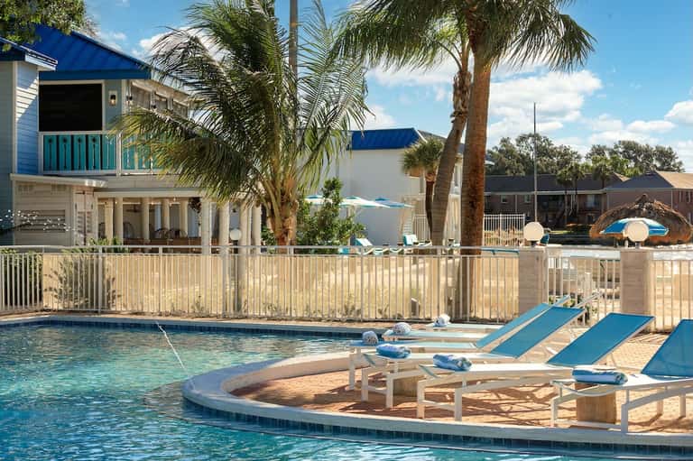 Beachfront resort pool with blue lounge chairs, spa area, palm trees, and elevated homes
