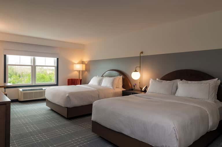 Double queen hotel room with white bedding, pendant lighting, windows, and warm color scheme