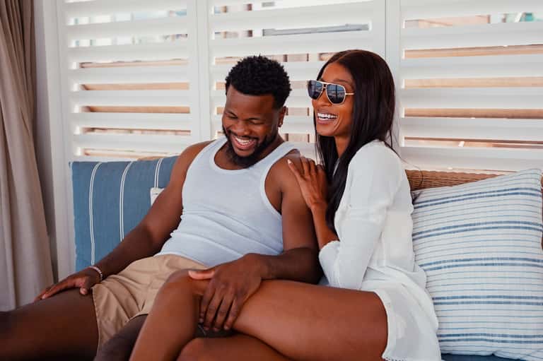 Couple sitting on bed in resort room with striped bedding and louvered window blinds