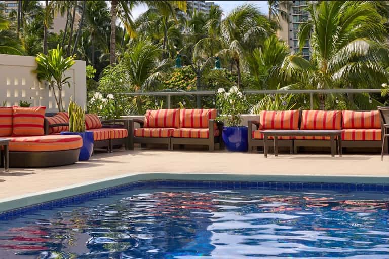 Resort pool with colorful striped lounge chairs, tropical vegetation, and tall hotel towers visible