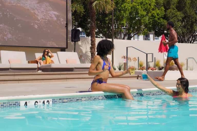 Modern pool area with guests swimming, white lounge seating, projection screen, and palm trees overhead